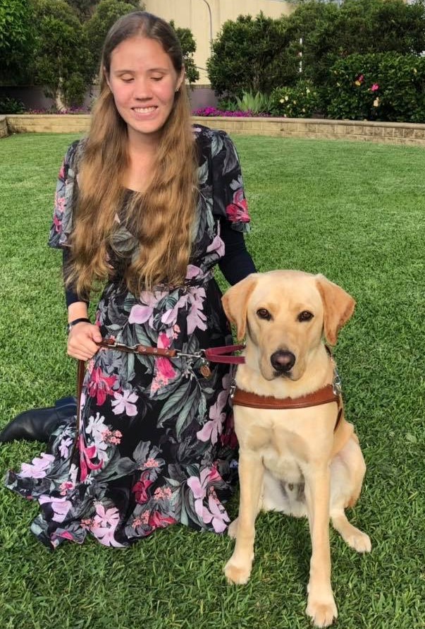Marianne and Zorro, a yellow labrador, are sitting on a well kept lawn in a garden. Zorro is wearing a guide dog harness and looking at the camera with an alert expression.