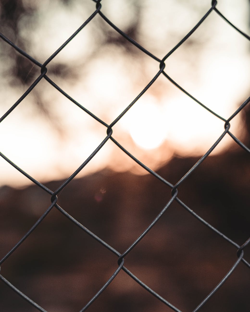 Image of a chain link fence with a blurred background. The image is used here to represent the experience of being kept out by a very visible barrier, without being able to even ascertaion what it is that is behind the barrier and cannot be accessed.